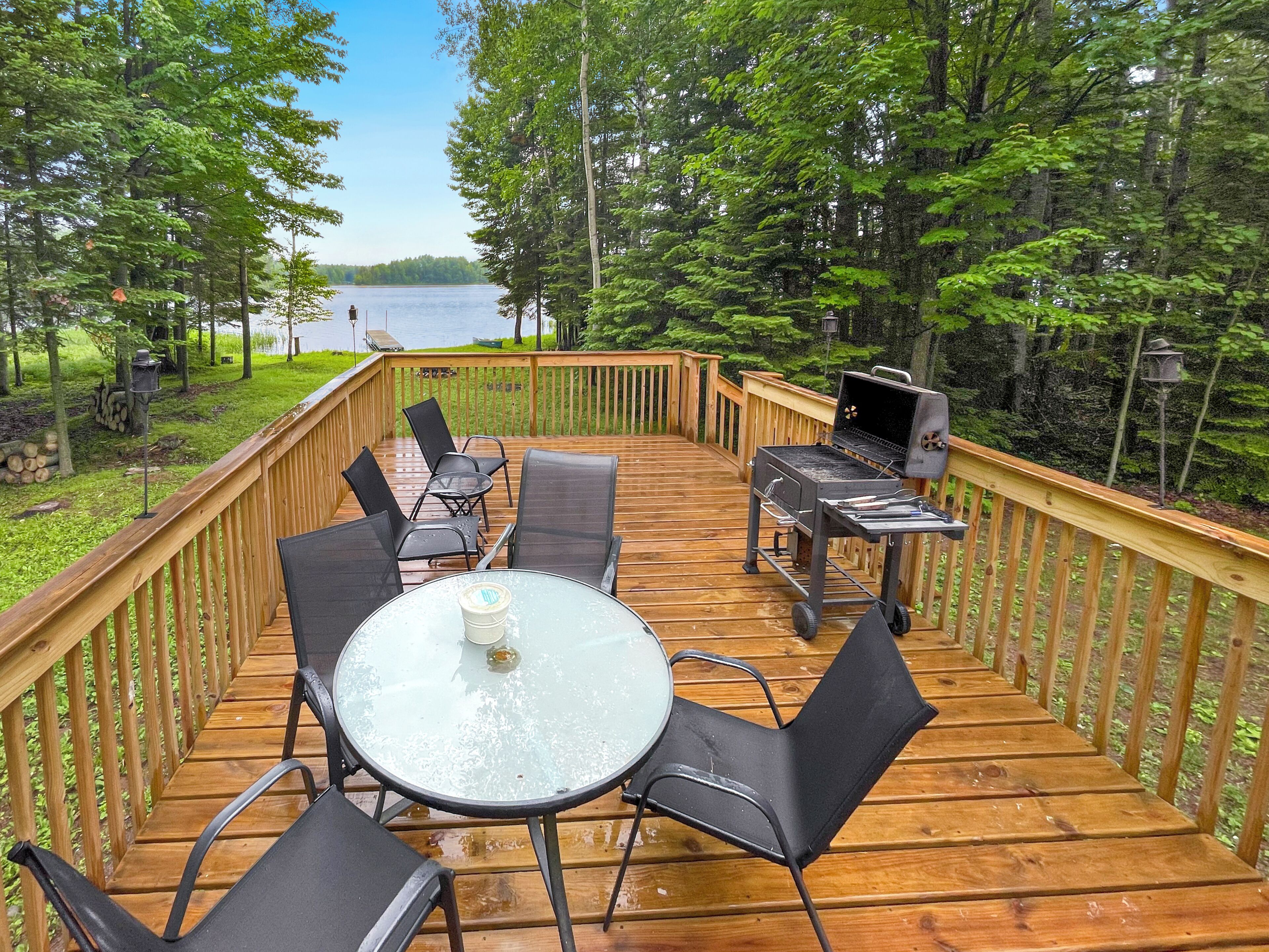 A wooden deck with a round glass table and chairs, a grill, and a view of a lake.