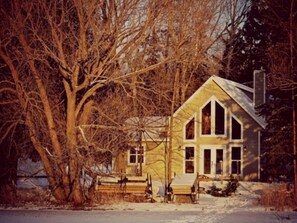 A two-story house with a gabled roof, surrounded by snow-covered ground and leafless trees.
