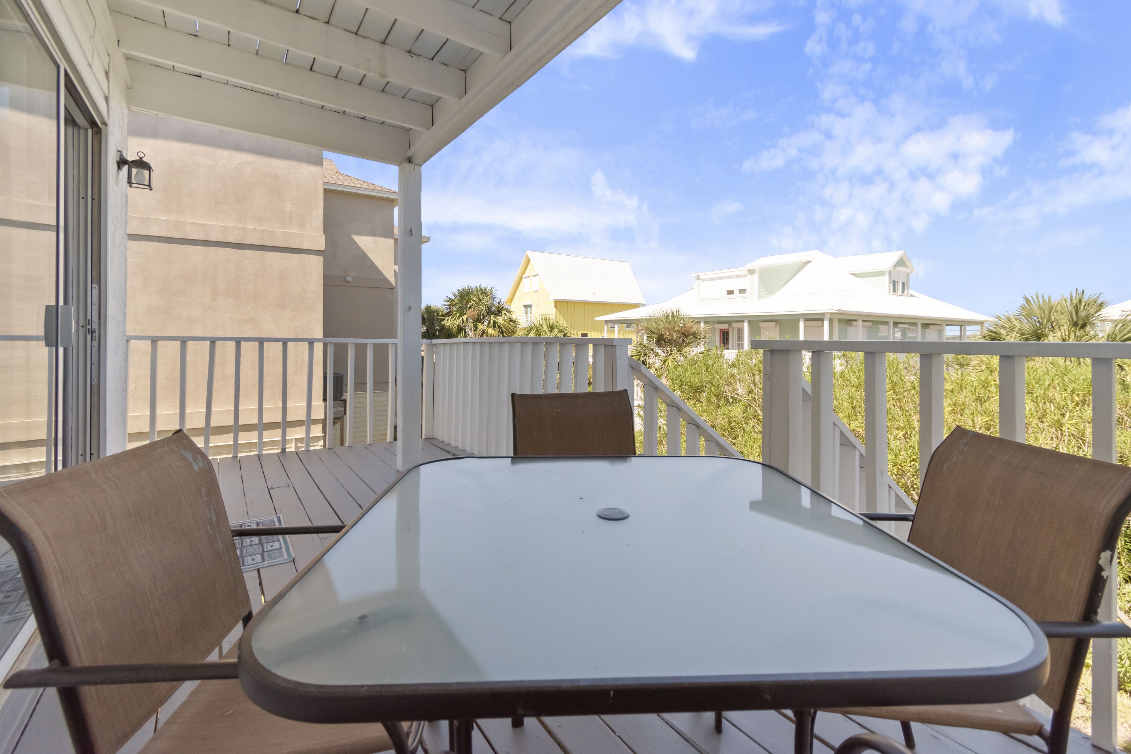 A covered outdoor patio with a glass table and two chairs, a white railing, and a view of houses in the background.