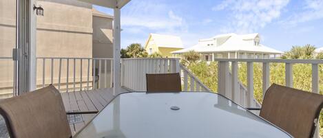 A covered outdoor patio with a glass table and two chairs, a white railing, and a view of houses in the background.