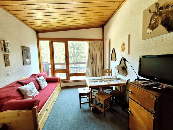 Een knus interieur met een houten plafond, een rode bank, een tafeltje, een televisie en een raam met gordijnen.