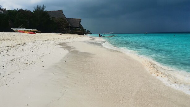 A beach with a thatched-roof structure, clear turquoise waters, and a cloudy sky. - Savi House Nungwi (Zanzibar)