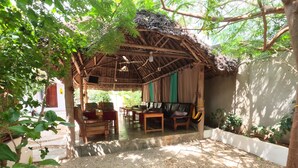 A covered outdoor seating area with a thatched roof, wooden furniture, and a gravel floor. - Savi House Nungwi (Zanzibar)