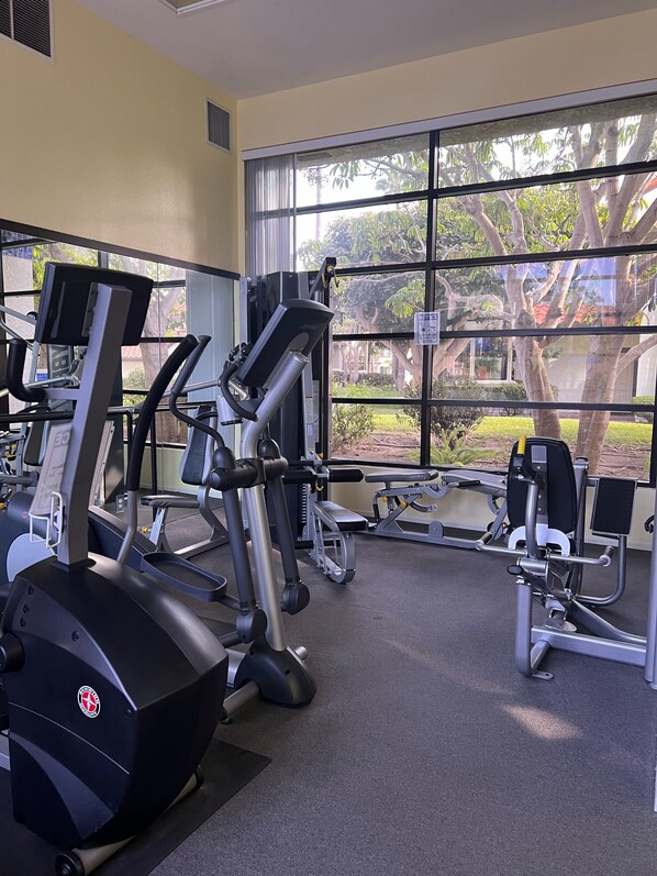 A gym with various exercise machines, large windows, and a view of trees and a building outside.