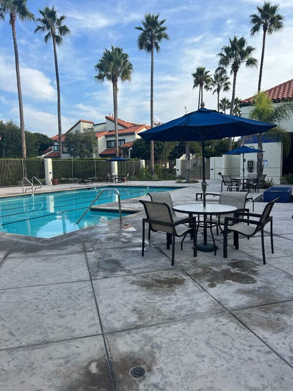 A pool area with a tiled deck, a table with chairs, and a blue umbrella. - Paradise at Mandalay Beach Resort living (Oxnard)