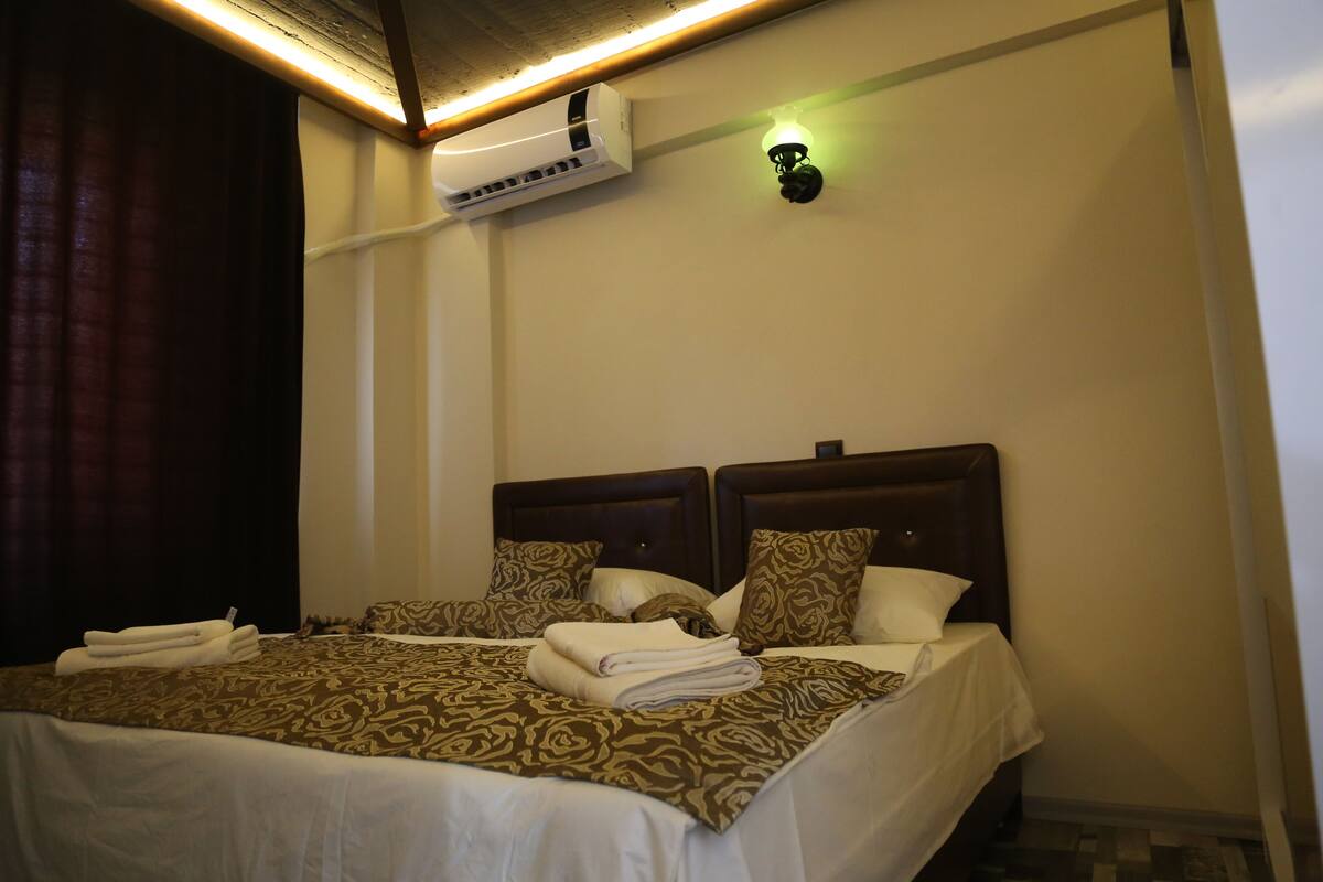 A hotel room with a large bed, two bedside tables, a wall-mounted air conditioner, and a wall sconce.