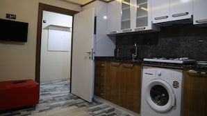 Una cocina compacta con armarios blancos, lavadora integrada y un otoman rojo.