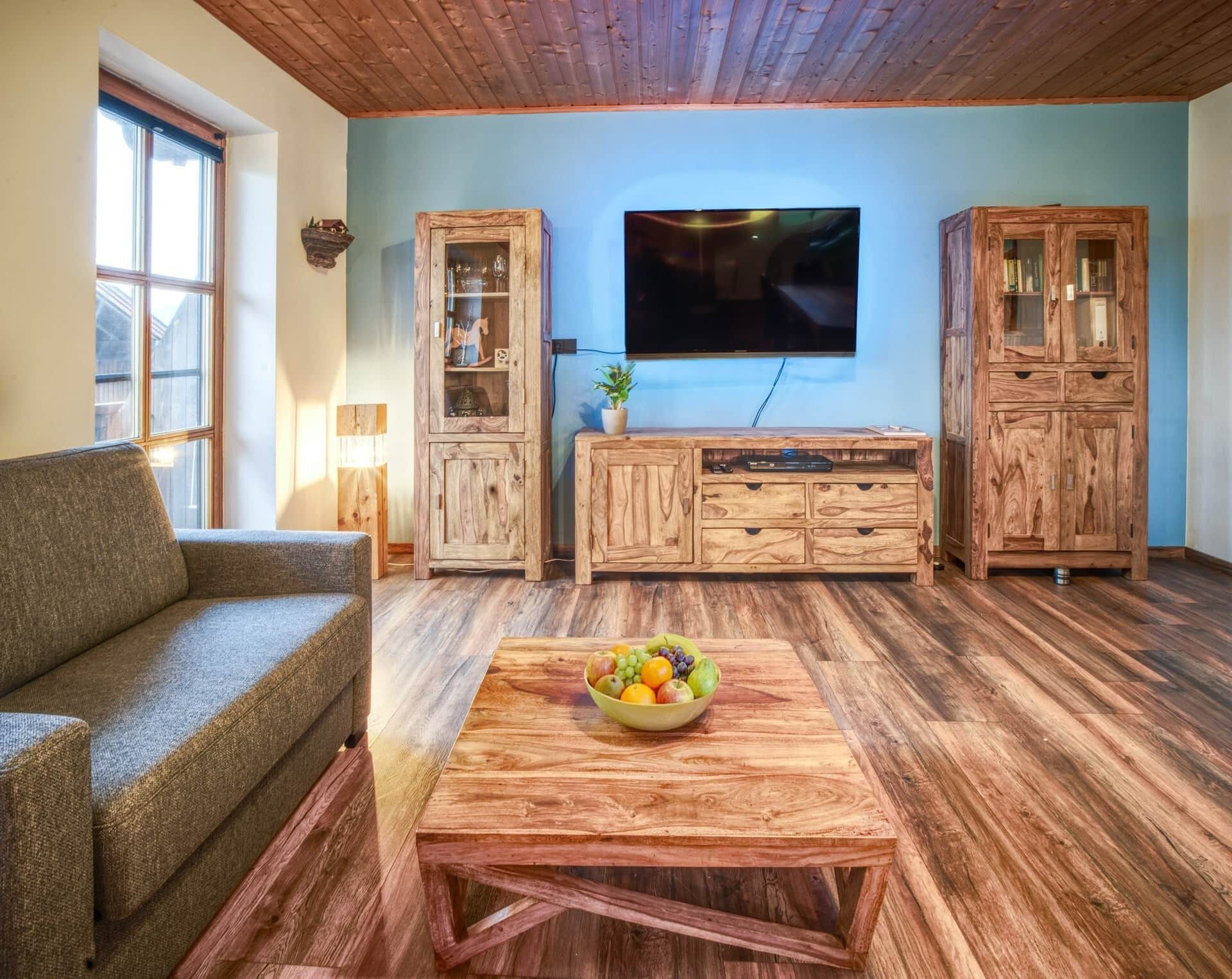 A living room with a wooden floor, a sofa, a coffee table with fruit, wooden cabinets, and a TV on the wall.