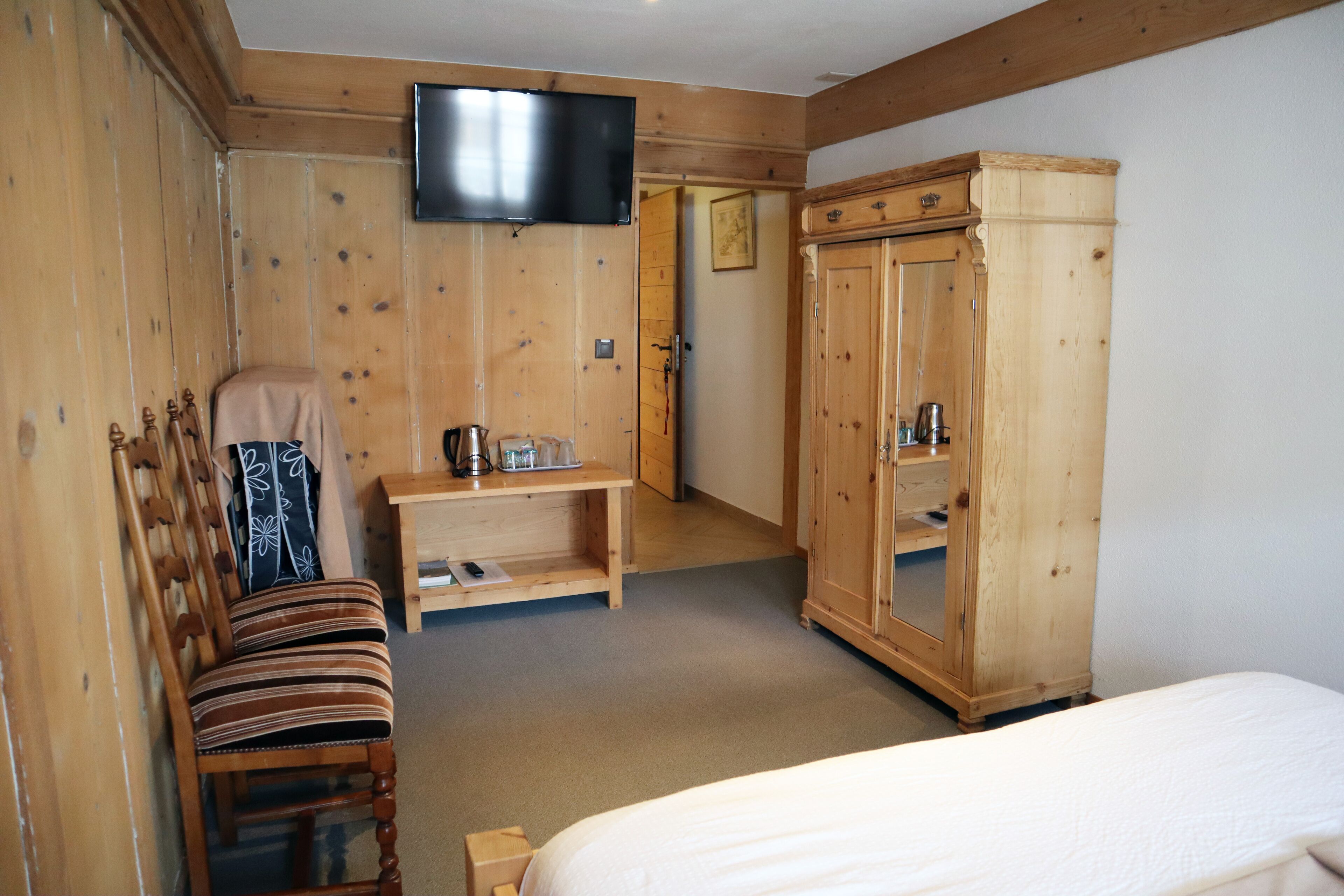 A wooden room with a bed, a wooden wardrobe, a television mounted on the wall, and a bench with a cushion.