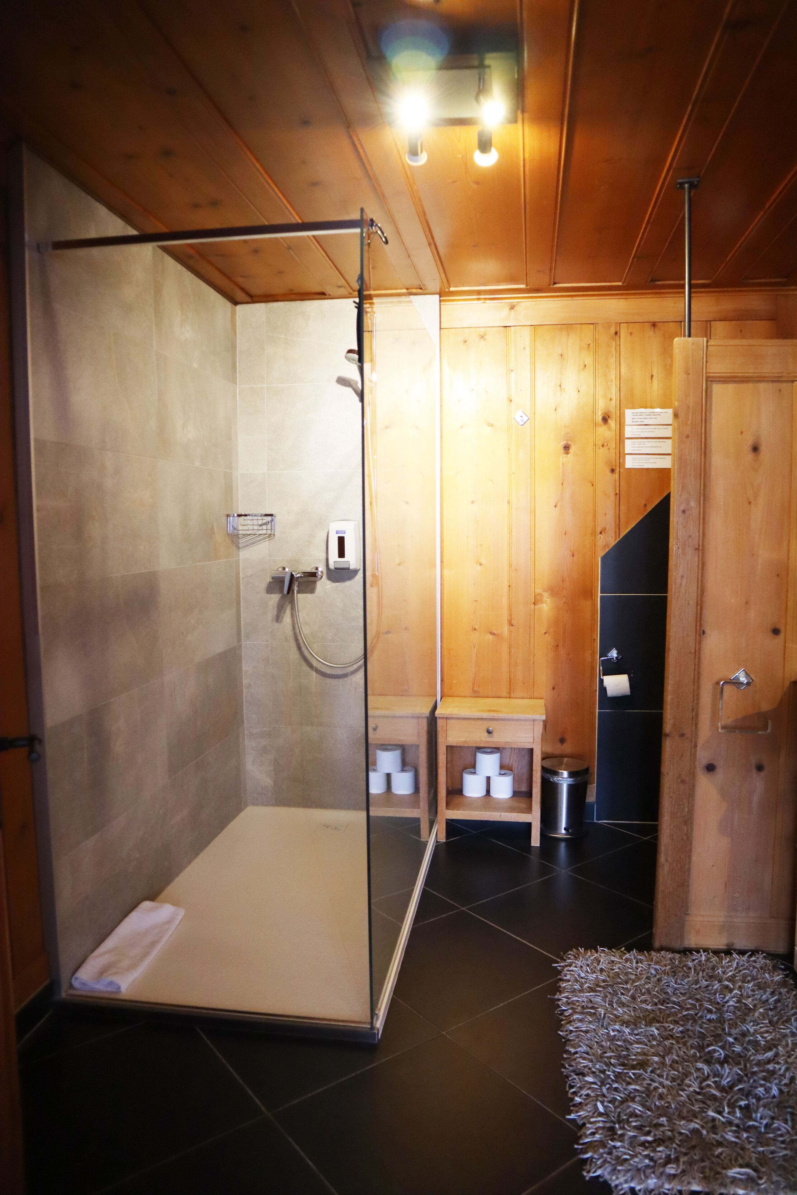 Une salle de bain rustique, avec une cabine de douche en verre, un banc en bois et un sol carrelé de carreaux noirs.
