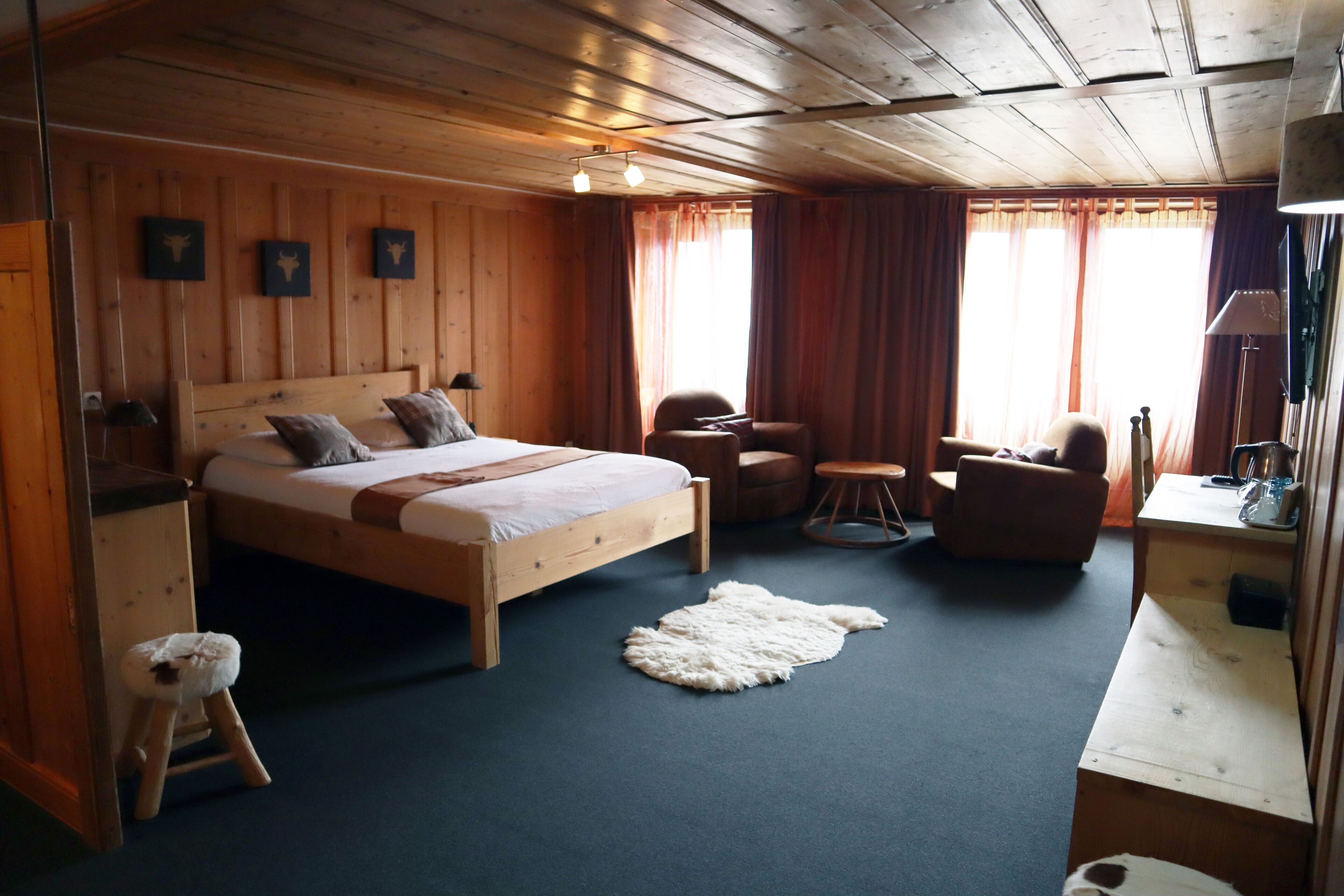 A wooden-paneled bedroom with a bed, two chairs, a small stool, a round table, and a desk with a lamp.