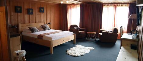 A wooden-paneled bedroom with a bed, two chairs, a small stool, a round table, and a desk with a lamp.