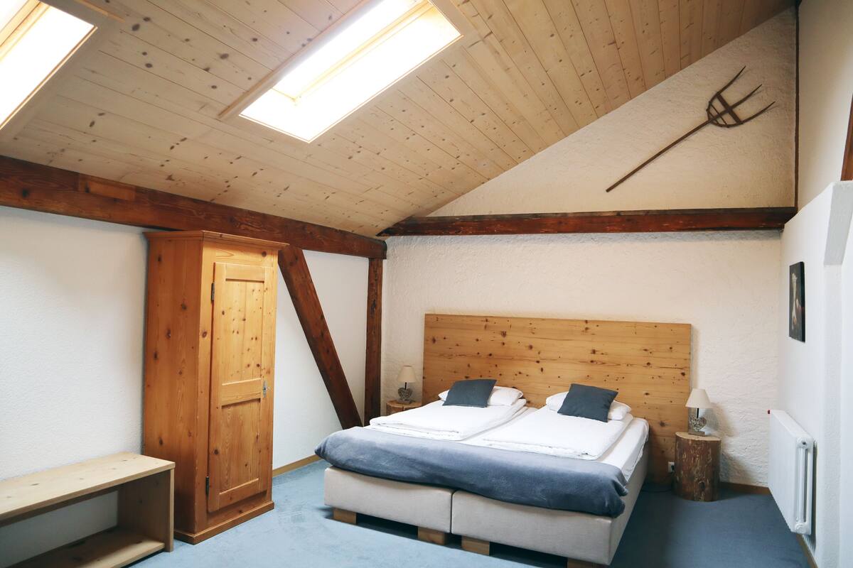 A bedroom with a sloped ceiling, wooden beams, a bed with two pillows, a bedside table with a lamp, and a wooden door.
