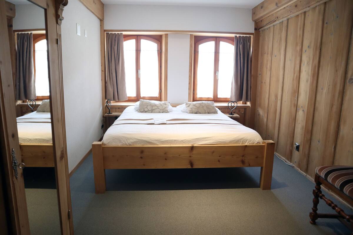A wooden-framed bed with a white mattress and two pillows, a wooden chair, and a window with curtains.