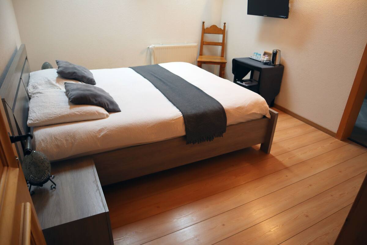 A neatly made bed with pillows and a blanket, a wooden chair, a small table with a lamp, and a television on the wall.