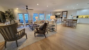 A spacious living room with a large window, a ceiling fan, and a dining area with a kitchen in the background.