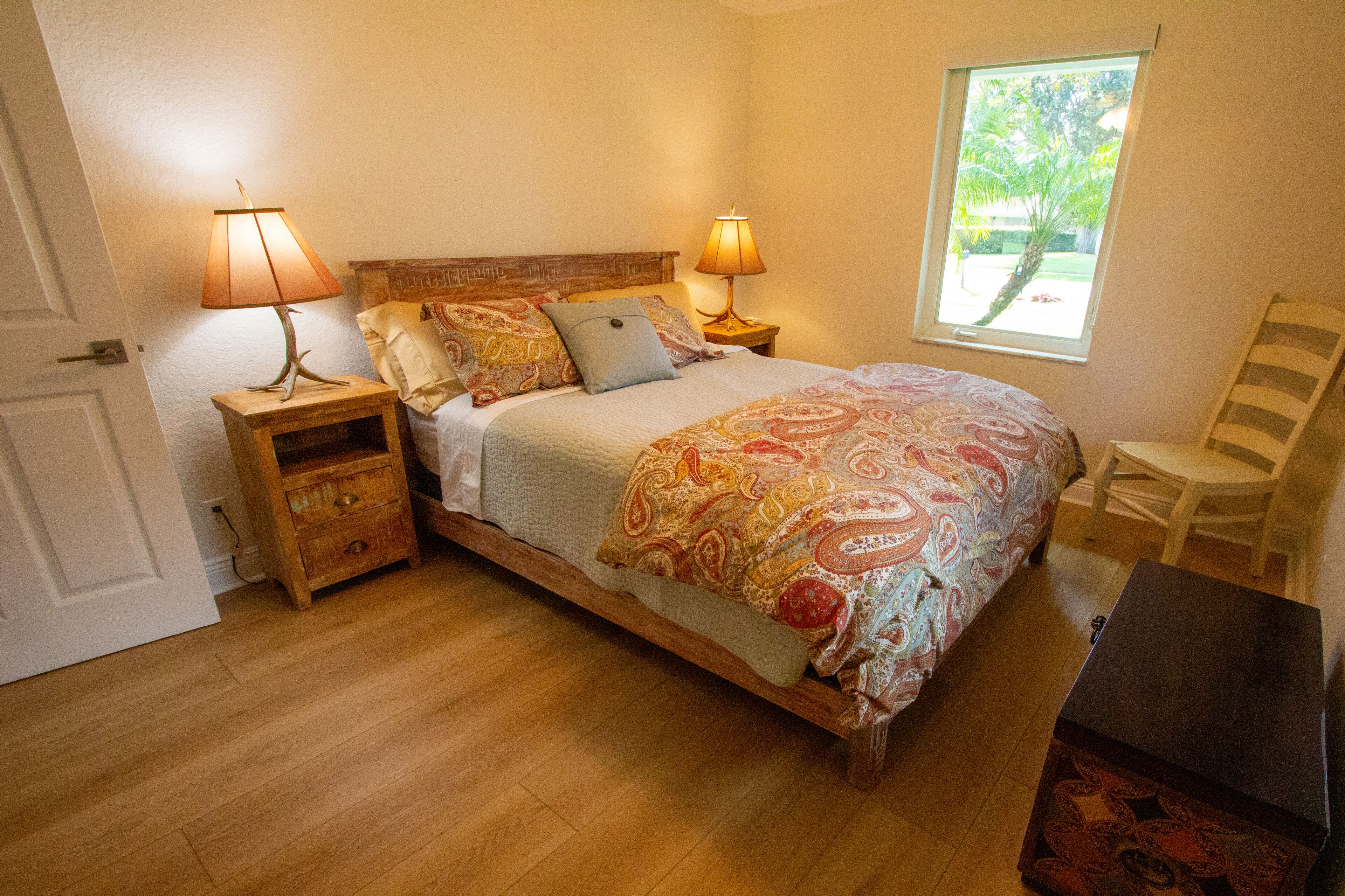 A bedroom with a wooden bed, two bedside tables with lamps, a chair, and a window with a view of greenery.