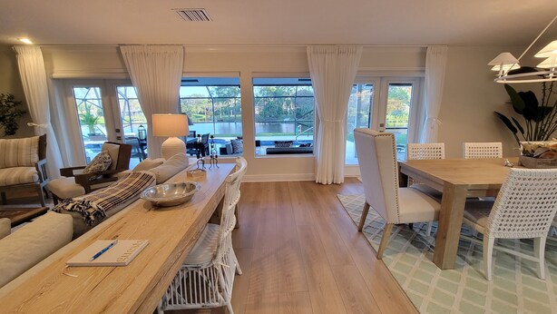 A spacious living area with a wooden dining table, white cushioned chairs, a sofa, and a large window offering a view of a pool and greenery.