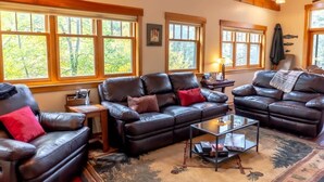 A cozy living room with leather sofas, a glass coffee table, and large windows offering a view of trees. - Tranquil 3BR Home; Access To Trails & Mtn. Views! (Breckenridge)