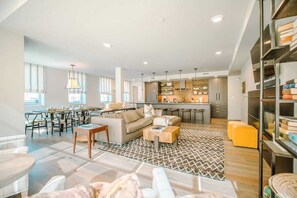 A spacious living area with a sofa set, coffee table, and dining tables. A kitchen with modern appliances and a bar area is visible in the background.