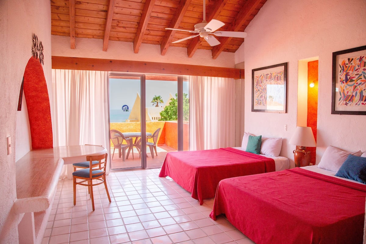 Una habitación con dos camas, una mesa de comedor, sillas, un ventilador de techo y vista a la playa.