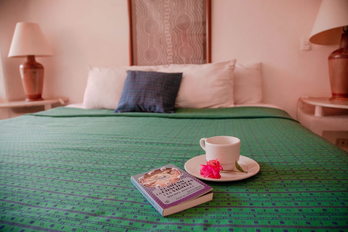 Una cama bien hecha con una colcha verde, un libro, una taza blanca y una flor rosa en un plato.