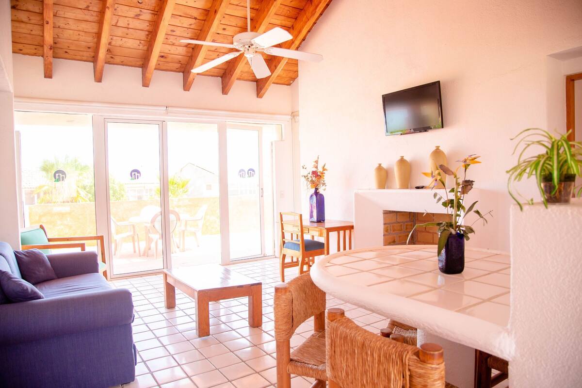 Una habitación con ventilador de techo, televisor montado en la pared, mesa de comedor con sillas, sofá y puerta corrediza de vidrio que da a un espacio al aire libre.