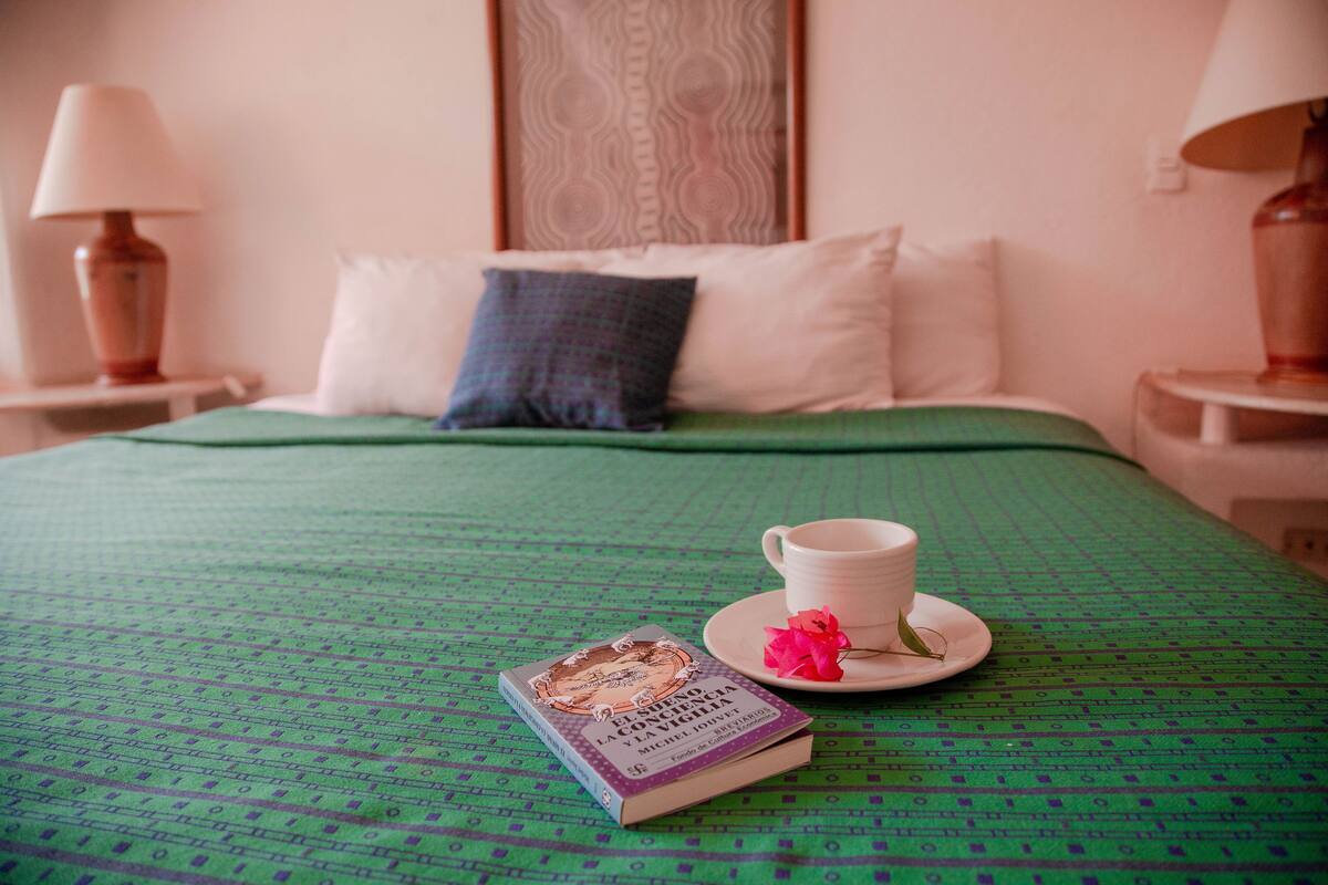 Una cama bien tendida con un edredón verde, un libro, una taza blanca y una flor rosa sobre un platillo a juego.