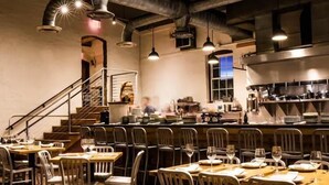 Una sala da pranzo con tavoli apparecchiati, una cucina sullo sfondo e una scala che conduce a un piano superiore.
