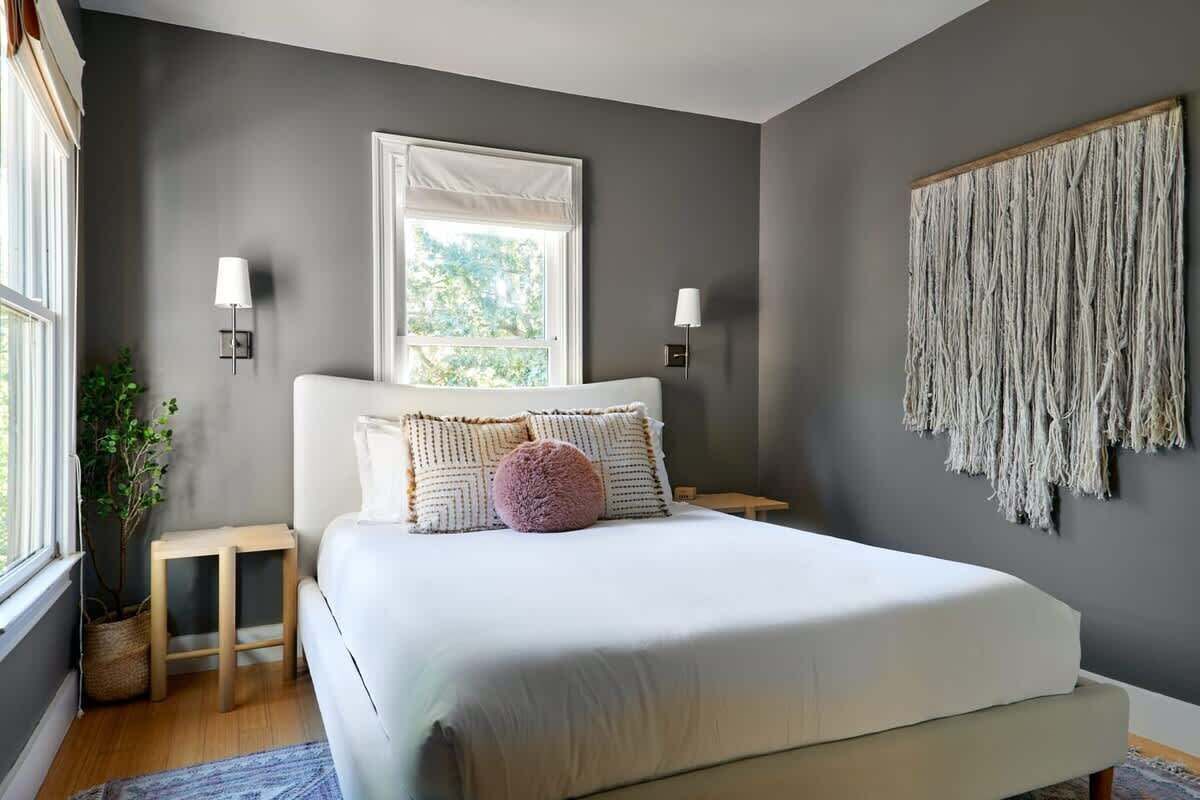 A bedroom with a large bed, two bedside tables, a window, and a wall-mounted macrame piece.