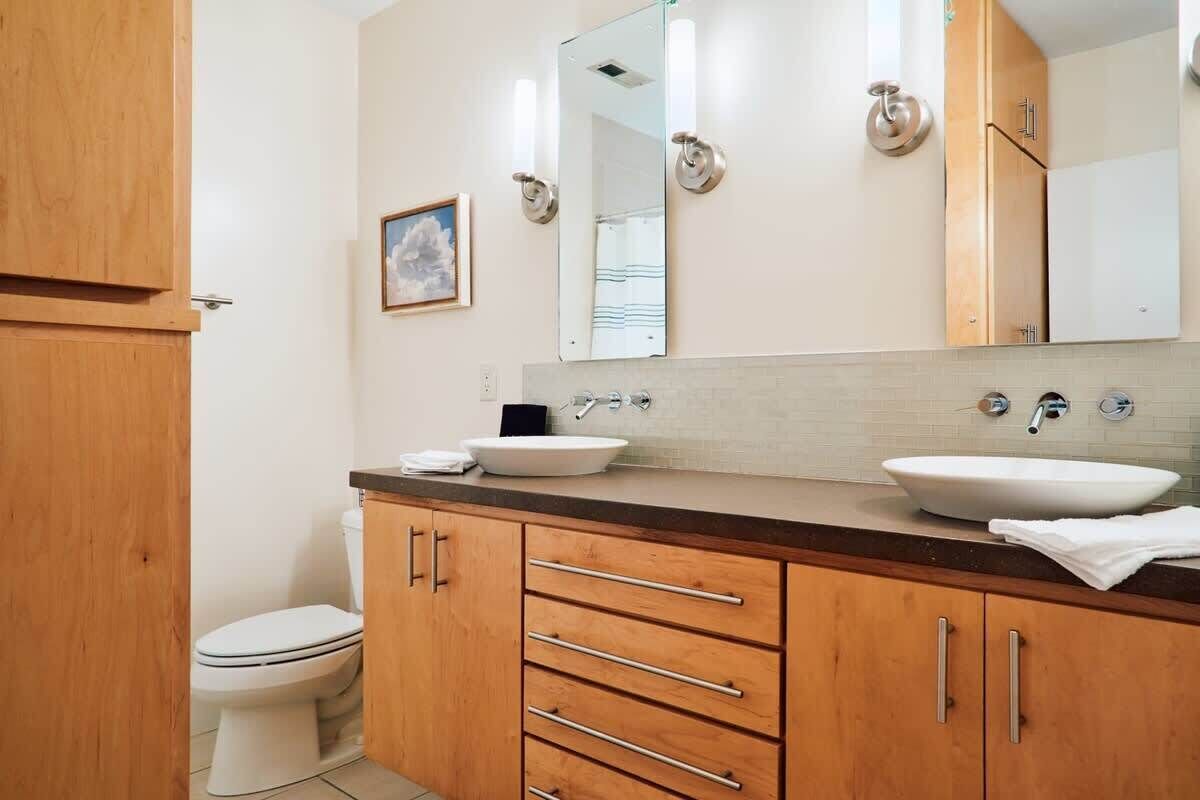 A modern bathroom with a wooden vanity, two sinks, a toilet, and a large mirror.
