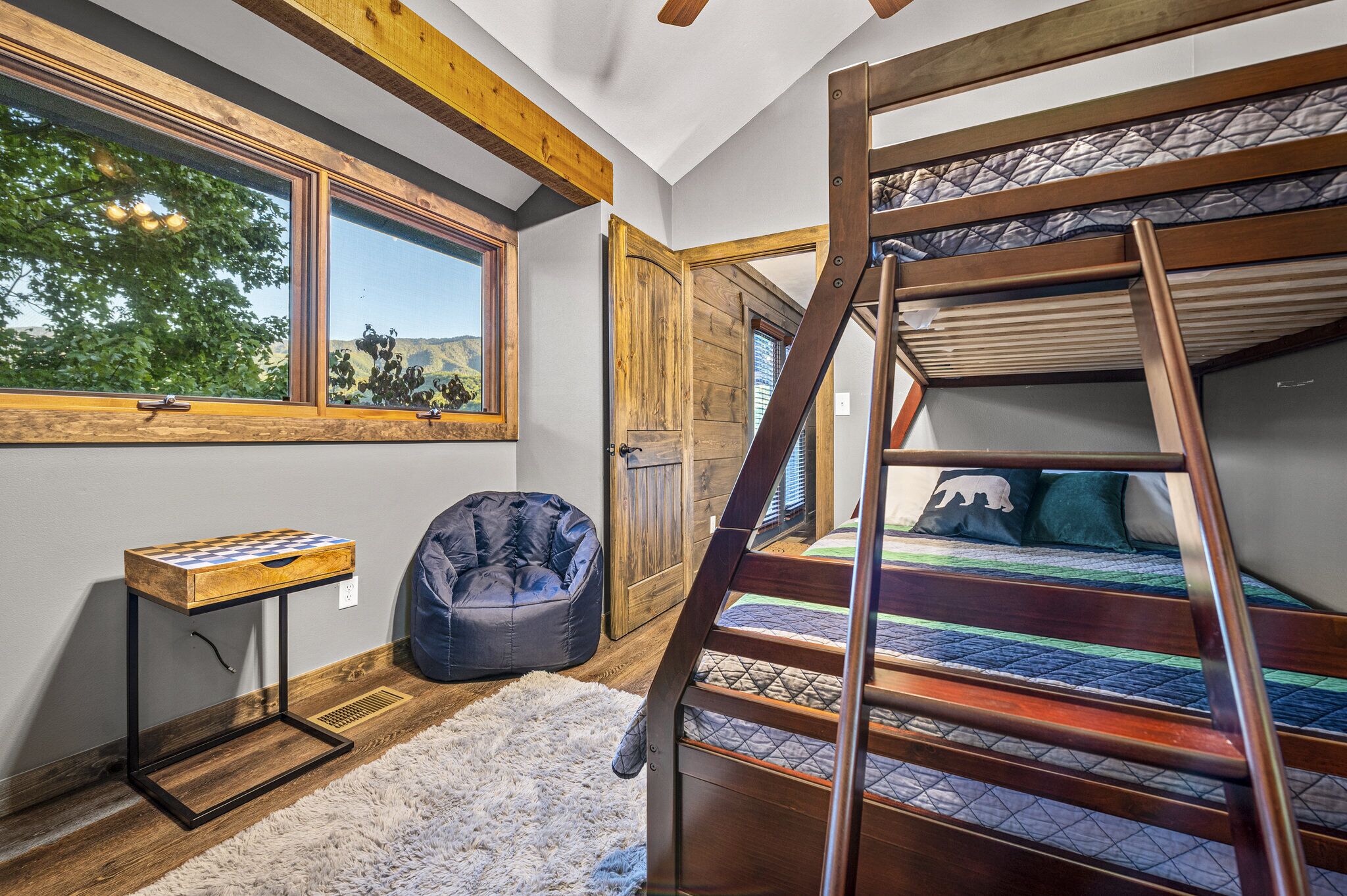 A room with a bunk bed, a desk with a board game, a bean bag chair, and a large window overlooking greenery.