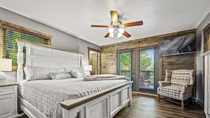 A bedroom with a large bed, a ceiling fan, wooden flooring, a plaid armchair, and a view of the outdoors through a sliding glass door. - THE Smoky Mountain Views, Comfort, Style, Games  (Gatlinburg)
