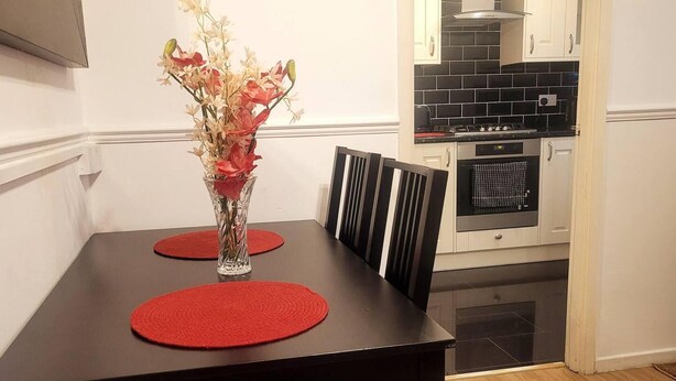 Une table à manger agrémentée d’un vase de fleurs, de sets de table rouges et de chaises noires, dans un coin cuisine doté d’un mur recouvert de carreaux et d’un four encastré.