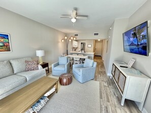 A modern living room with a sofa, armchairs, a coffee table, and a TV. There is a kitchen area in the background with a dining table and chairs.