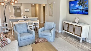 Ein Wohnzimmer mit zwei Sesseln, einem Couchtisch, einer Essecke mit Tisch und Stühlen, einer weißen Kommode mit Fernseher und einer gerahmten Karte an der Wand.