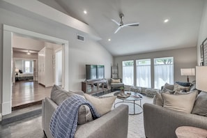 A spacious living room with a high ceiling, a ceiling fan, a flat-screen TV, a fireplace, and a large window with curtains. - Modern Luxurious Home-w/game room  (Fredericksburg)