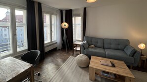 Sala moderna com sofá cinzento, mesa de centro de madeira e abajur. Janelas amplas oferecem vista para edifícios.