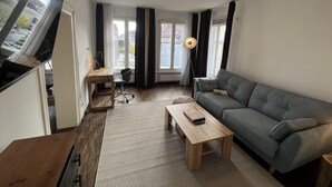Uma sala de estar moderna com um sofá, uma mesa de centro de madeira e uma escrivaninha com cadeira. Há um abajur e uma janela com cortinas.