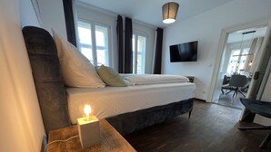 Quarto moderno com cama, mesa de cabeceira com abajur, televisão de tela plana e uma área de refeições visível por uma porta aberta.