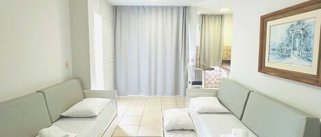 Habitación de hotel con dos camas individuales, un sofá, una mesita y un ventilador de techo, además de una ventana con cortinas.