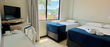 A hotel room with a desk, two beds, a TV, and a window with a view of palm trees and buildings.