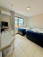 Quarto de hotel com escrivaninha, duas camas, televisão e janela com vista para palmeiras e edifícios.
