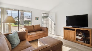 Een moderne woonkamer met een bruine leren bank, een houten tv-meubel en een groot raam met uitzicht op palmbomen en bergen.