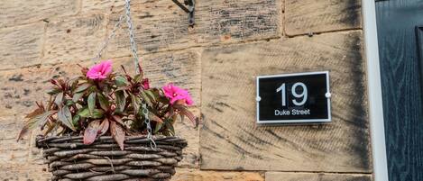 A stone wall with a hanging flower basket and a house number sign.