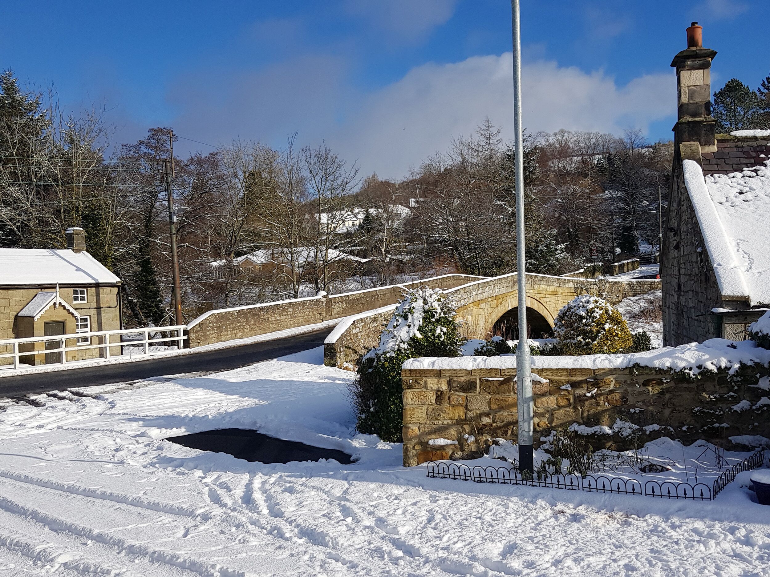 A stone bridge over a snow-covered road with a stone building on the left and a chimney on the right.