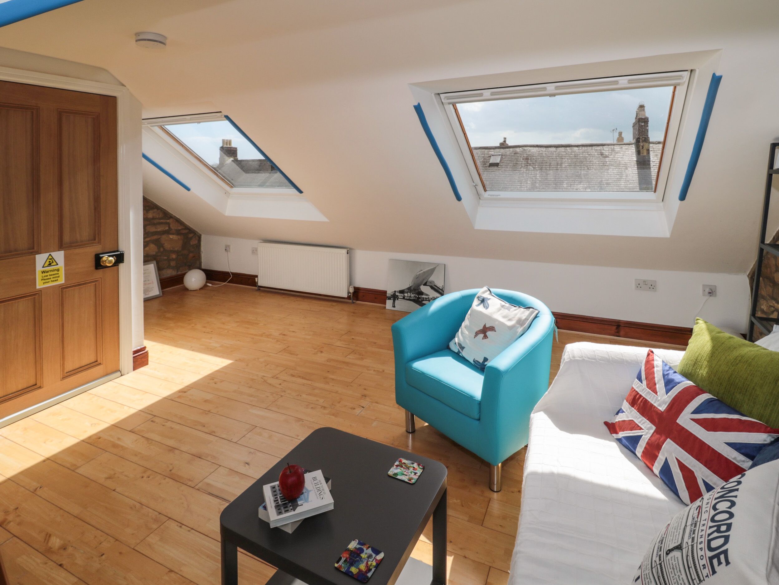 A room with a wooden floor, a blue armchair, a bed with a Union Jack pillow, and a skylight.