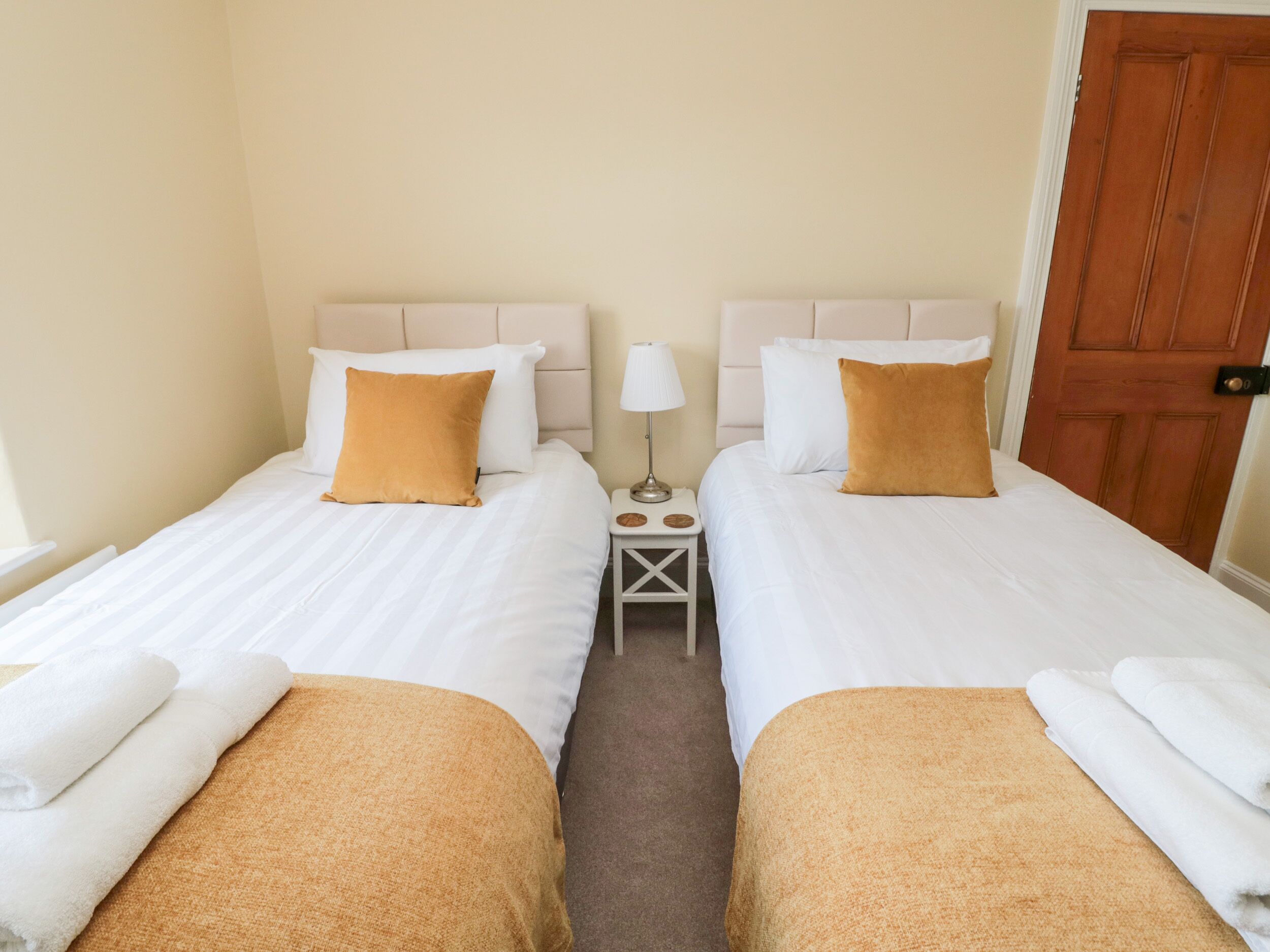 Une chambre d’hôtel avec deux lits, chacun agrémenté de draps blancs et d’oreillers décoratifs marron. On y trouve une petite table de chevet avec une lampe, ainsi qu’une commode avec une autre lampe. La chambre a une porte en bois et des murs de couleur beige.
