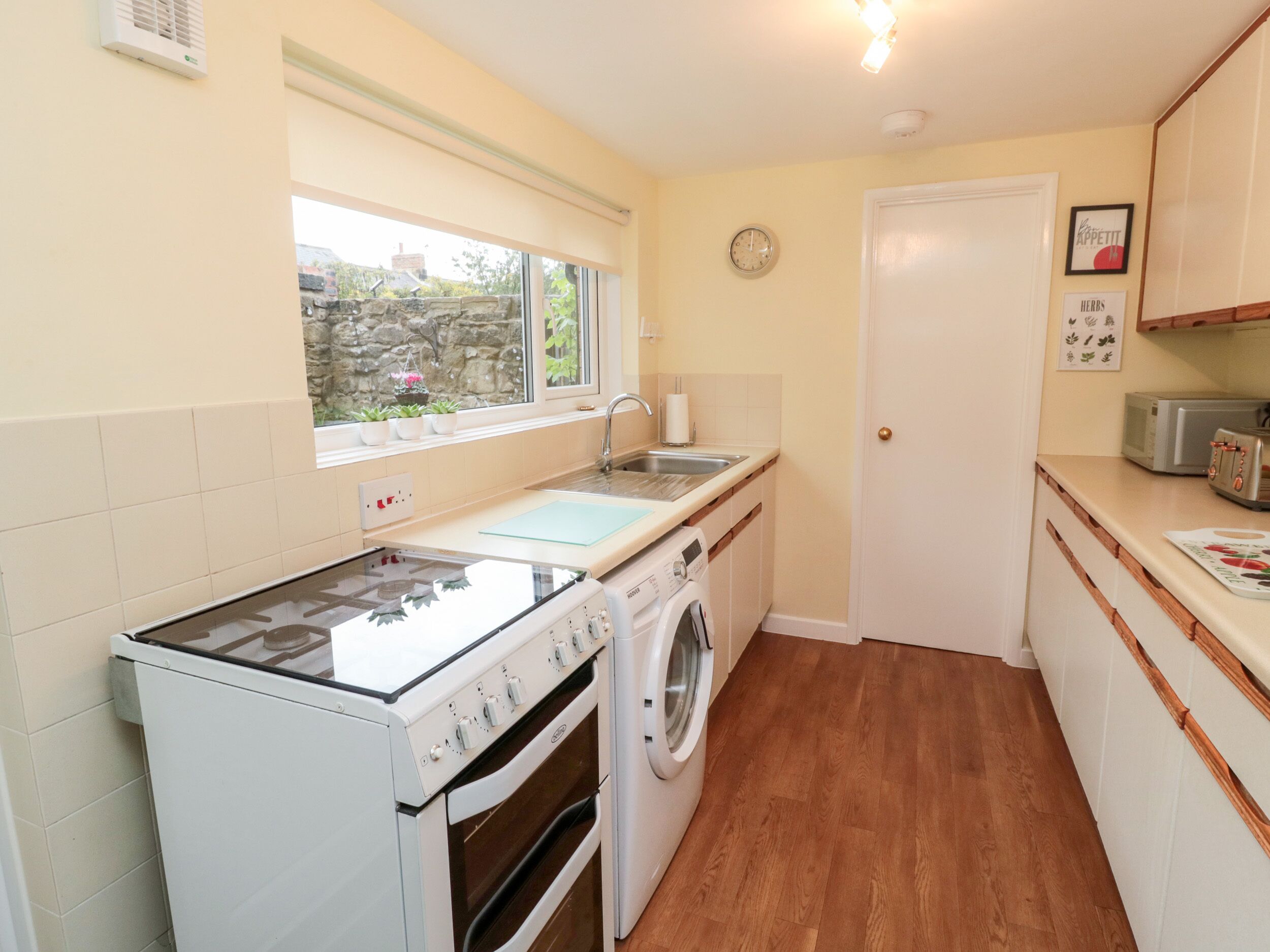 A compact kitchen with a stove, oven, washing machine, sink, and a window overlooking a stone wall.