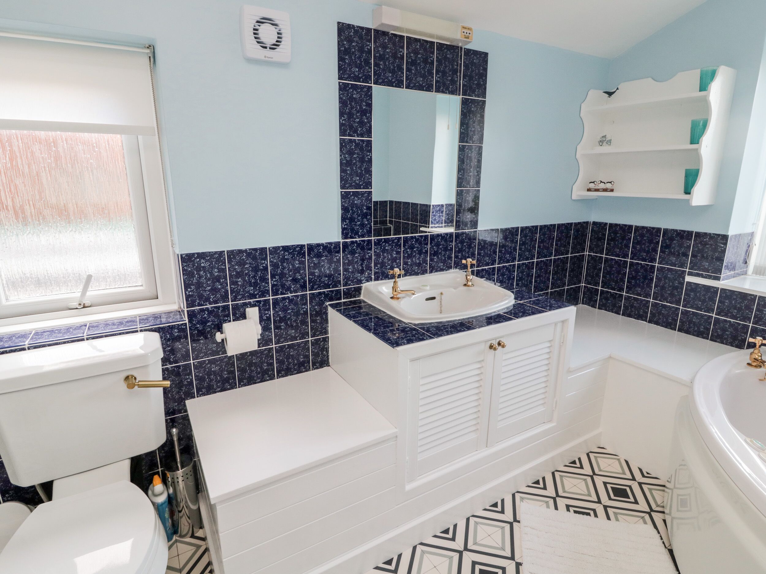 Une salle de bain avec des toilettes blanches, un lavabo avec des robinets dorés, une baignoire et un carrelage décoré.